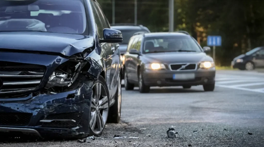 Ciemnoniebieski samochód z mocno uszkodzoną przednią lewą stroną jest zaparkowany na poboczu drogi. Na chodniku leżą porozrzucane odłamki. Za nim przejeżdżają dwa samochody na wielopasmowej drodze, z drzewami i znakami ulicznymi widocznymi w tle.