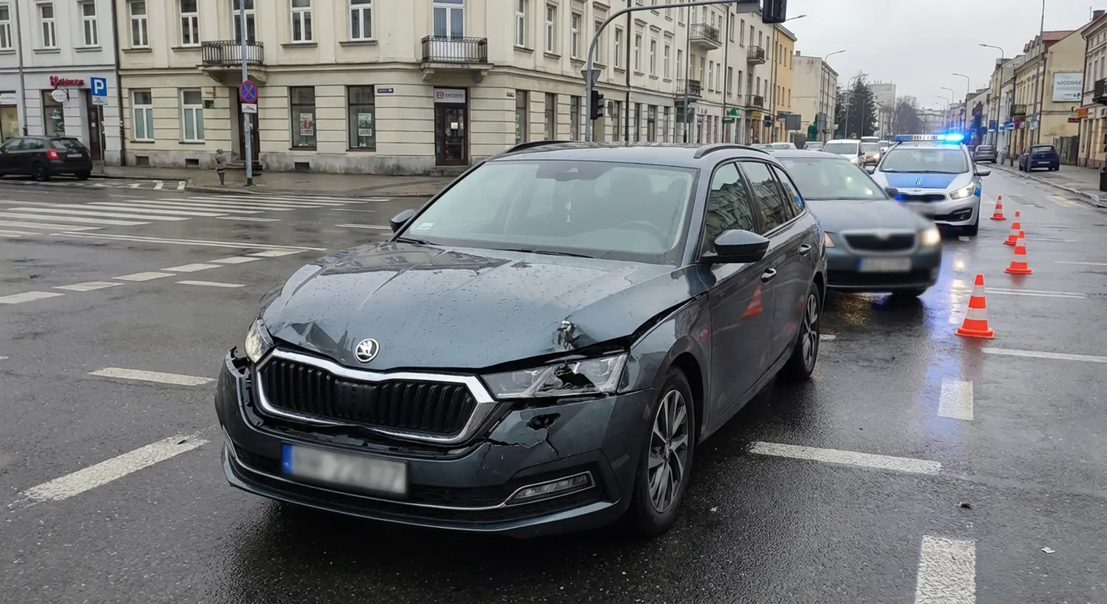 Ciemnoszary samochód z widocznymi uszkodzeniami przodu jest zatrzymany na mokrej ulicy miasta. Za nim stoją samochody policyjne z migającymi światłami i pachołkami drogowymi. Budynki i przejścia dla pieszych ciągną się wzdłuż ulicy pod zachmurzonym niebem.