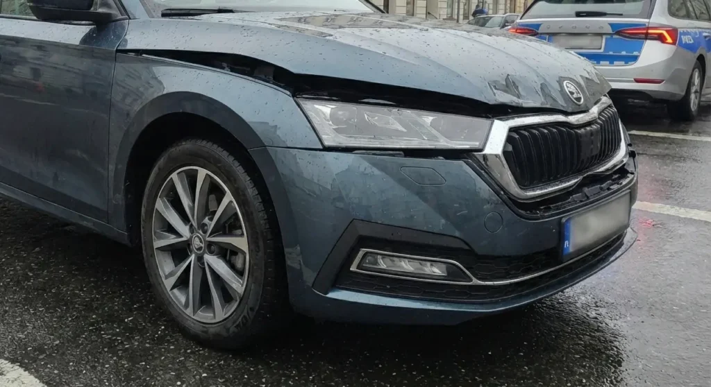 Niebieski samochód z uszkodzoną, pogniecioną maską i przednim zderzakiem jest zaparkowany na mokrej ulicy. Na pojeździe widoczny jest deszcz. W tle widać inny samochód, prawdopodobnie policyjny. Scena sugeruje niedawny wypadek.